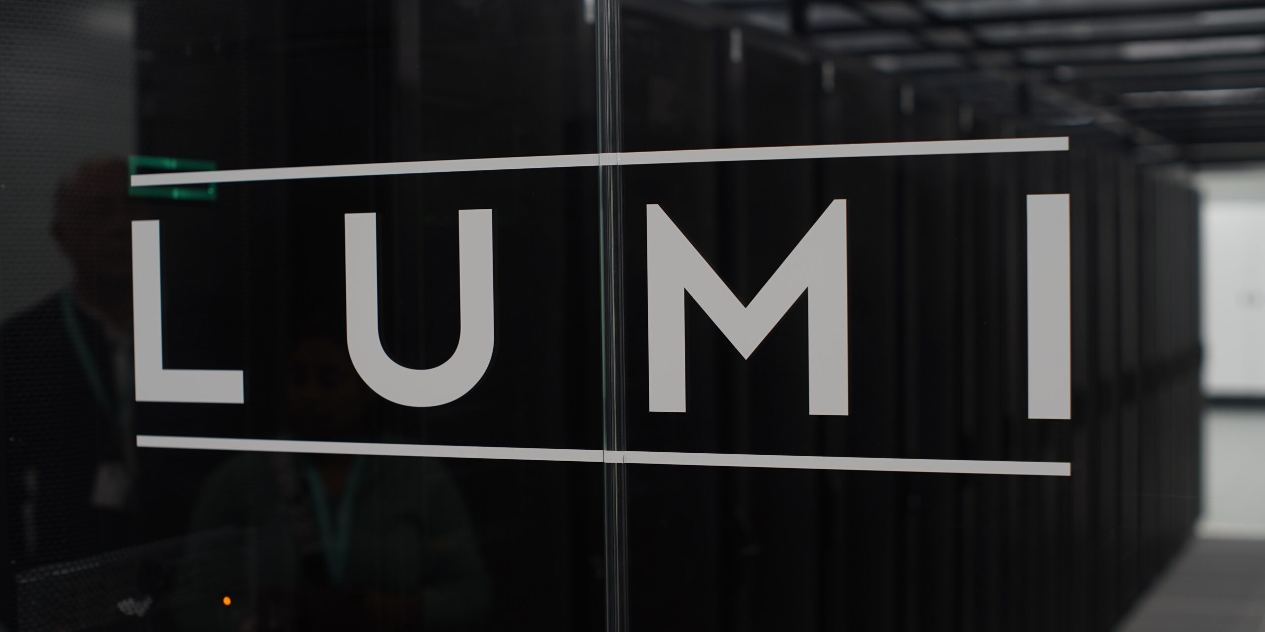 the word LUMI on a glass door. Behind we can see a supercomputer